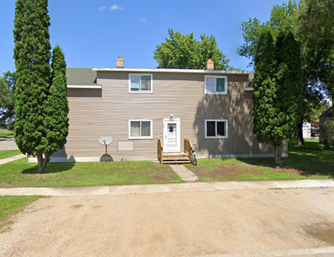 A house with a brown siding and a white door is surrounded by trees.