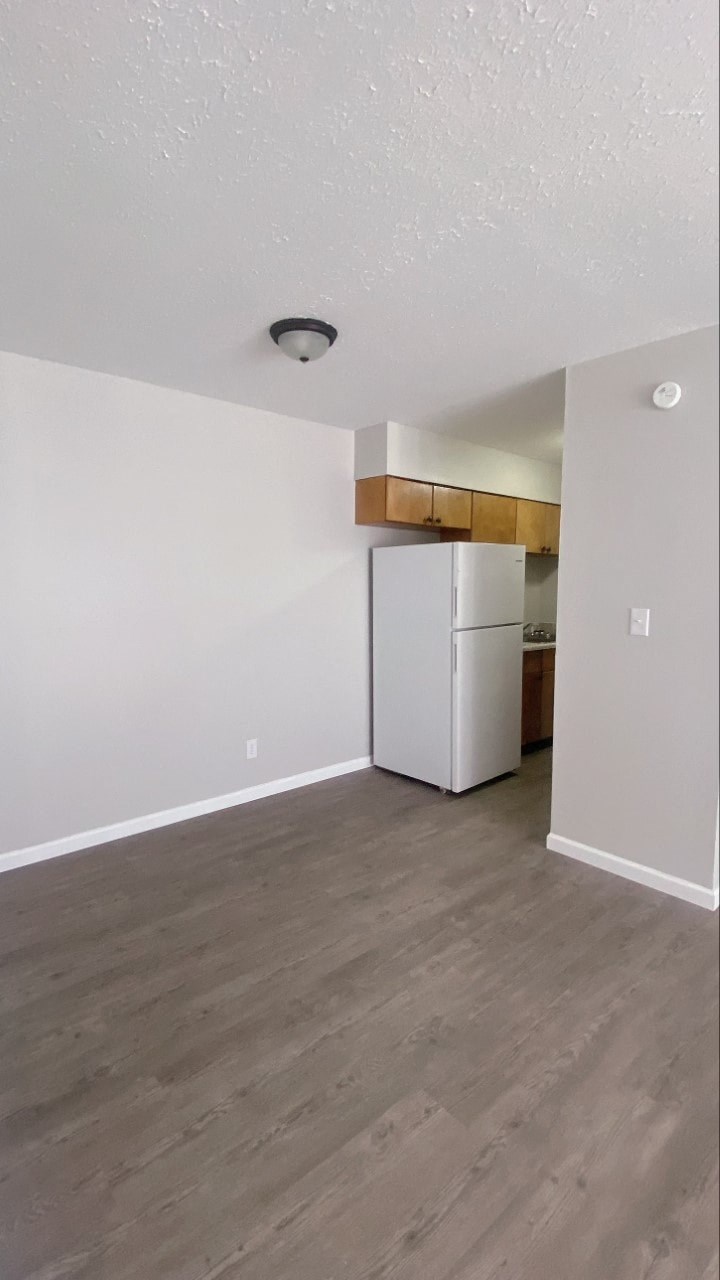 an empty living room with a refrigerator and a kitchen
