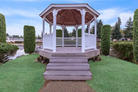 A gazebo with a wooden roof and white railings is surrounded by a grassy area and a paved walkway leading up to it.