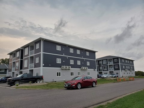 A red car is parked in front of a grey apartment building.