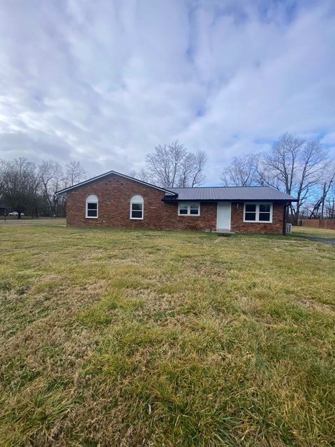 A small brick house with a white door and two windows sits in a grassy field.