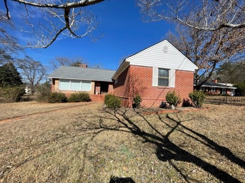 A house with a red brick exterior and a white roof is surrounded by a dry grassy area.