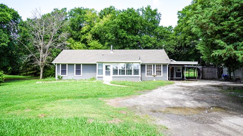 A house with a grey roof and white walls is surrounded by green grass and trees.
