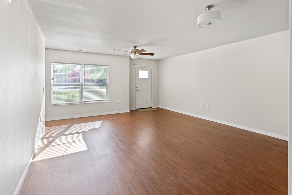 A spacious living room with wood-style flooring, large windows with natural light, and a ceiling fan at Muskego School Apartments