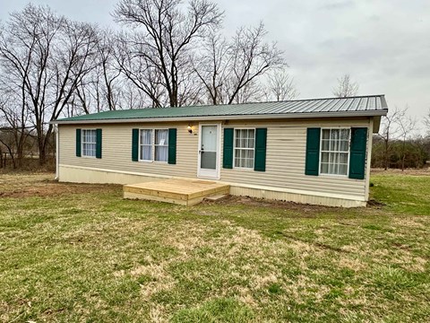 a small house with green shutters and a deck in a grassy field