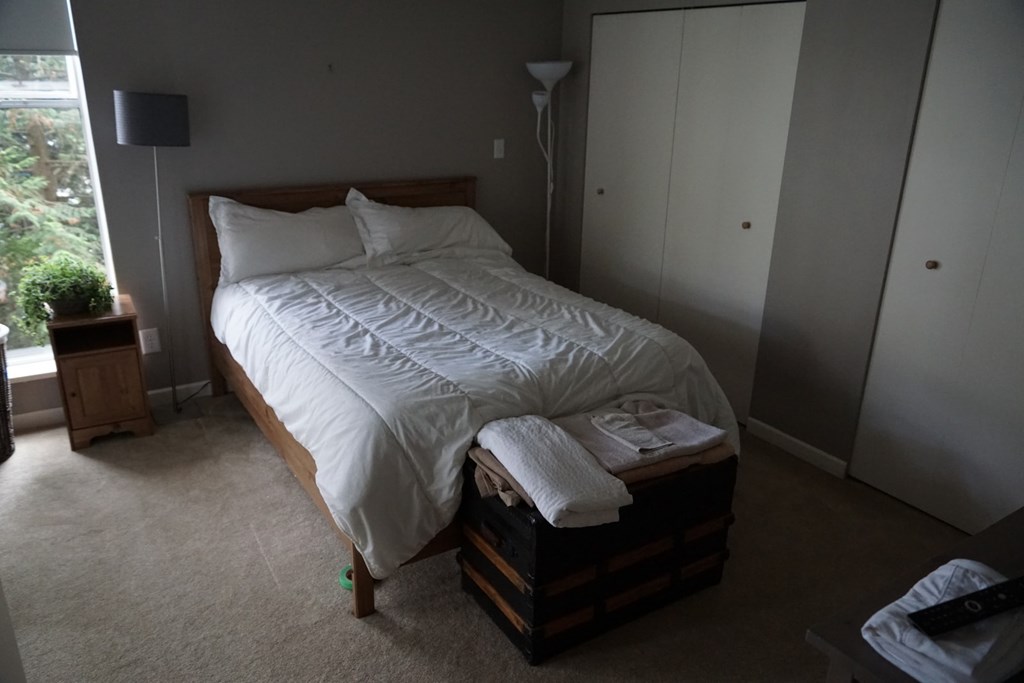 A bed with a white comforter and pillows sits in a room with a lamp and a window.