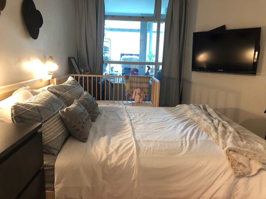 A bed with a white comforter and a grey pillow is in the foreground of a room with a window and a flat screen TV mounted on the wall.