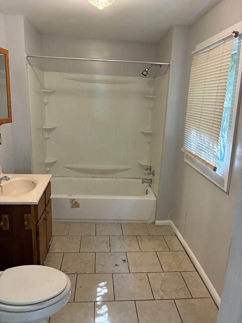 A white toilet is in a bathroom with a white tub and a window with blinds.