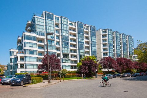 A person riding a bicycle in front of a large building with a parking lot in front.
