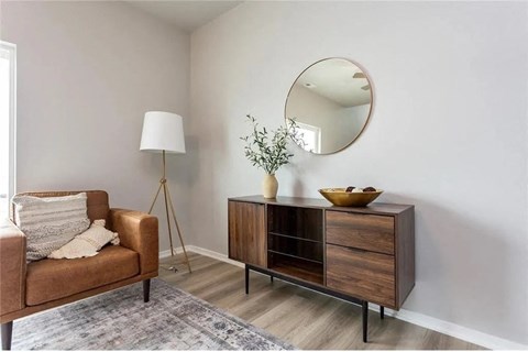 A brown chair sits next to a wooden cabinet with a round mirror above it.