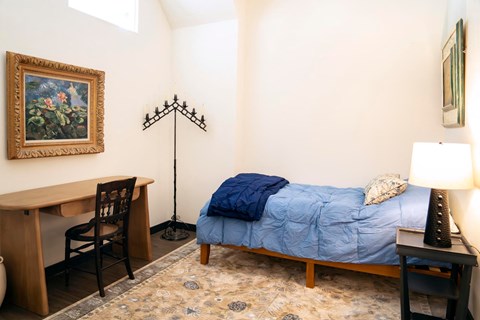 A bedroom with a bed, desk, chair, and a painting on the wall.