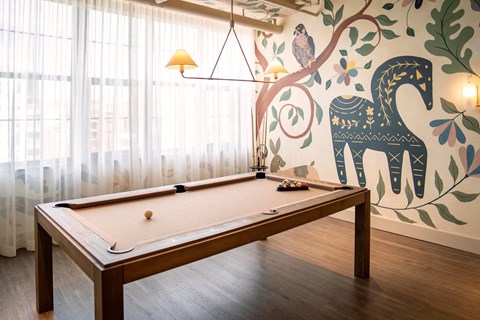 A pool table with a wooden frame and a green felt cloth is in the foreground of a room with a mural of a horse and trees on the wall.