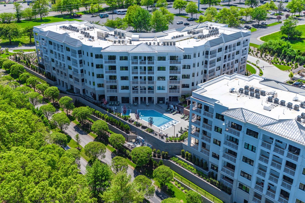 A large white hotel with a pool in the middle of a green area.