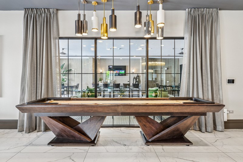 A large wooden table with a glass top is in the center of a room with grey curtains.