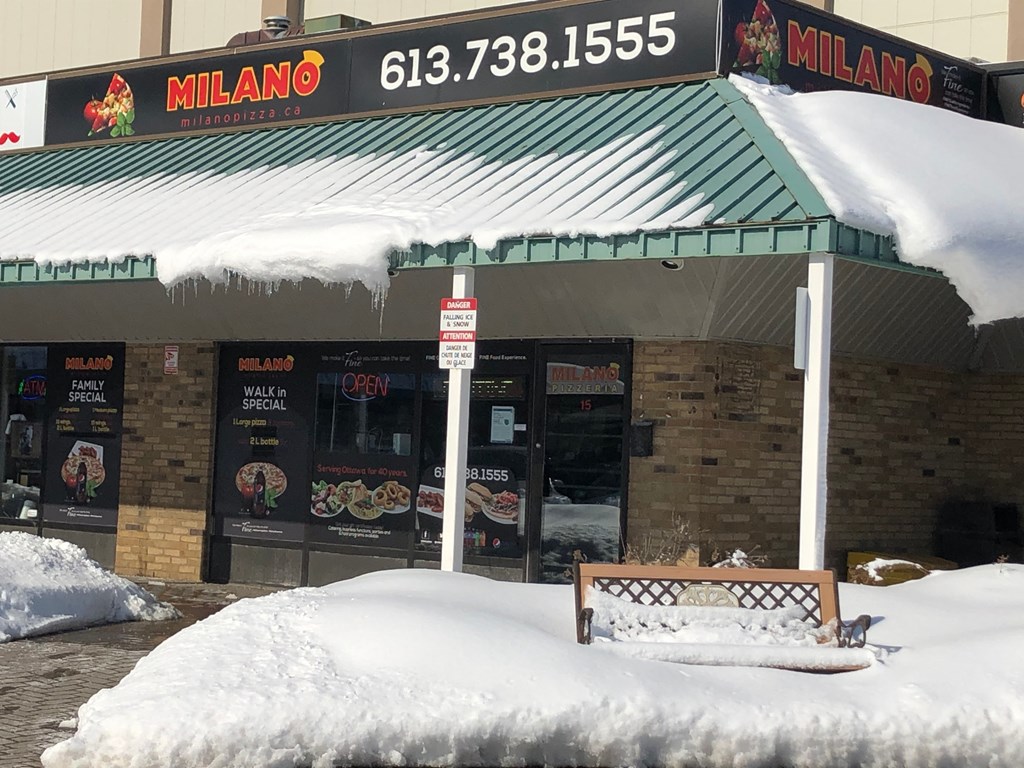 a milano restaurant covered in snow with a bench in front of it