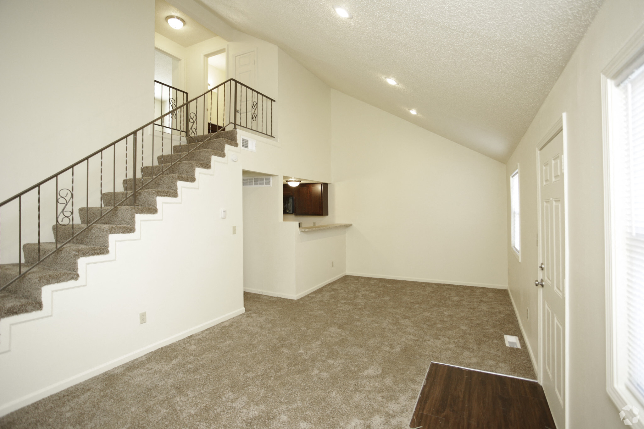 Spacious living room with white walls and gray carpeting with a view of a flight of stairs that lead to the loft  and a view of the kitchen with black appliances