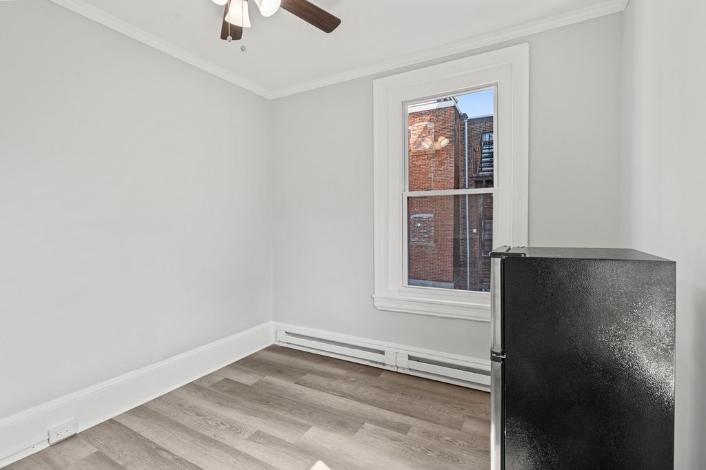 A black fridge sits in a room with a window and a ceiling fan.