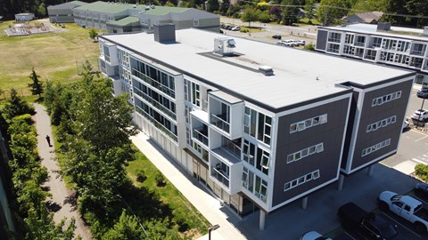 A modern building with a grey and white exterior is surrounded by greenery and other buildings.