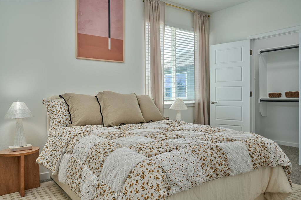 A bed with a floral patterned comforter and two pillows sits in a room with a lamp and a painting on the wall.