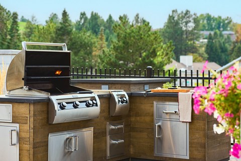 A BBQ grill is on a wooden deck with a black fence and flowers in the background.
