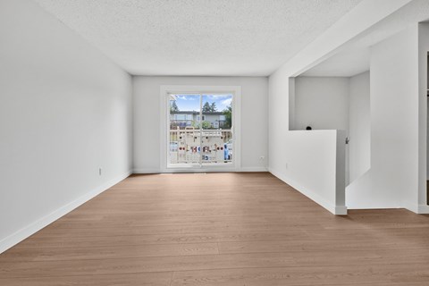 A room with wooden floors and white walls, with a large window that shows a view of a street with cars and buildings.