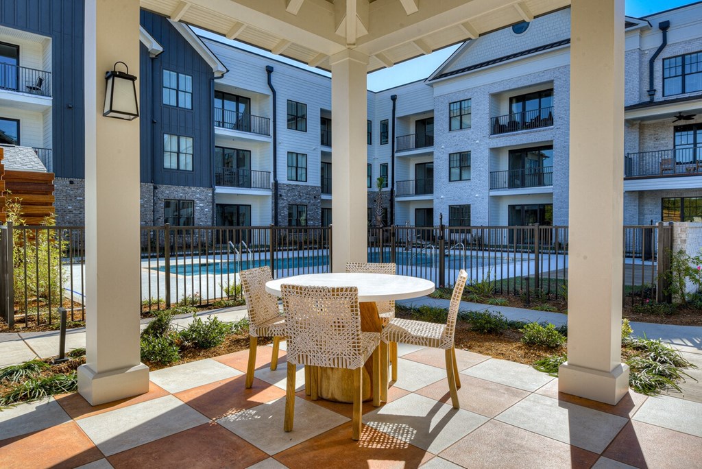 A patio with a table and chairs is surrounded by a pool and apartment buildings.