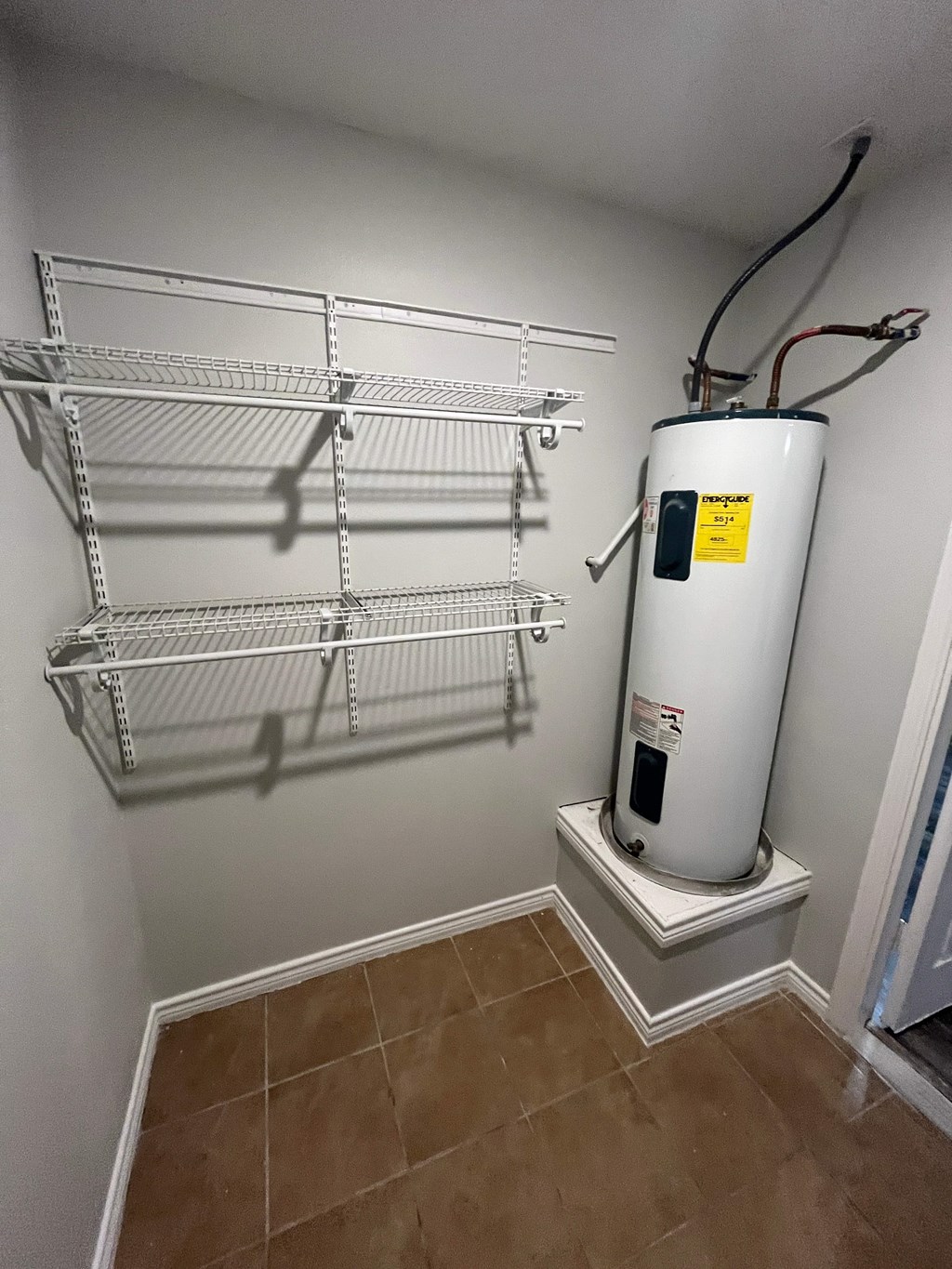 A water heater is installed in a corner of a room with a metal shelf above it.