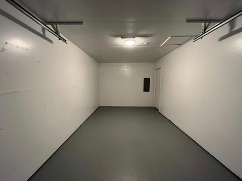 A long, narrow, white-walled room with a grey floor and a door at the end.