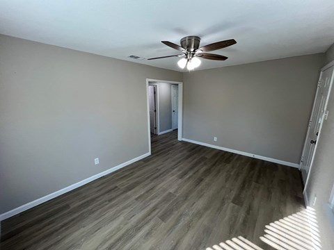 A room with a ceiling fan and light fixture, hardwood floors, and a doorway leading to another room.