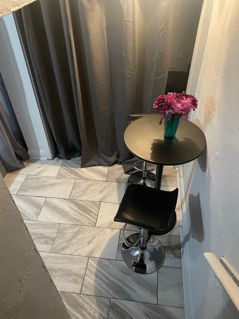 A black stool is in front of a round table with a vase of pink flowers on it.