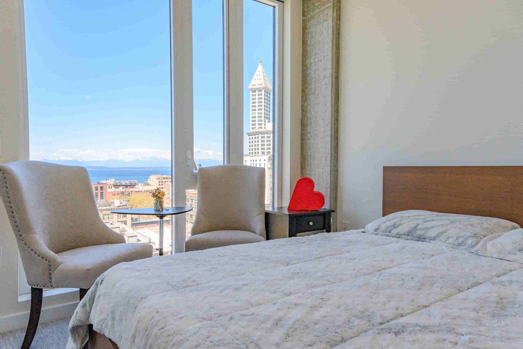 A bedroom with a large bed, a chair, and a view of a cityscape through a window.