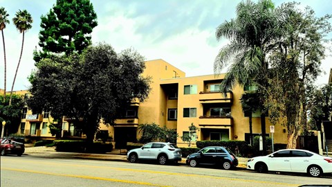 A building with a yellow facade is surrounded by trees and cars are parked in front.