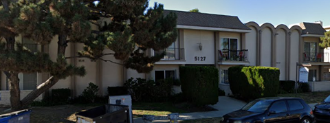 A black car is parked in front of a two-story building with a balcony on the second floor.