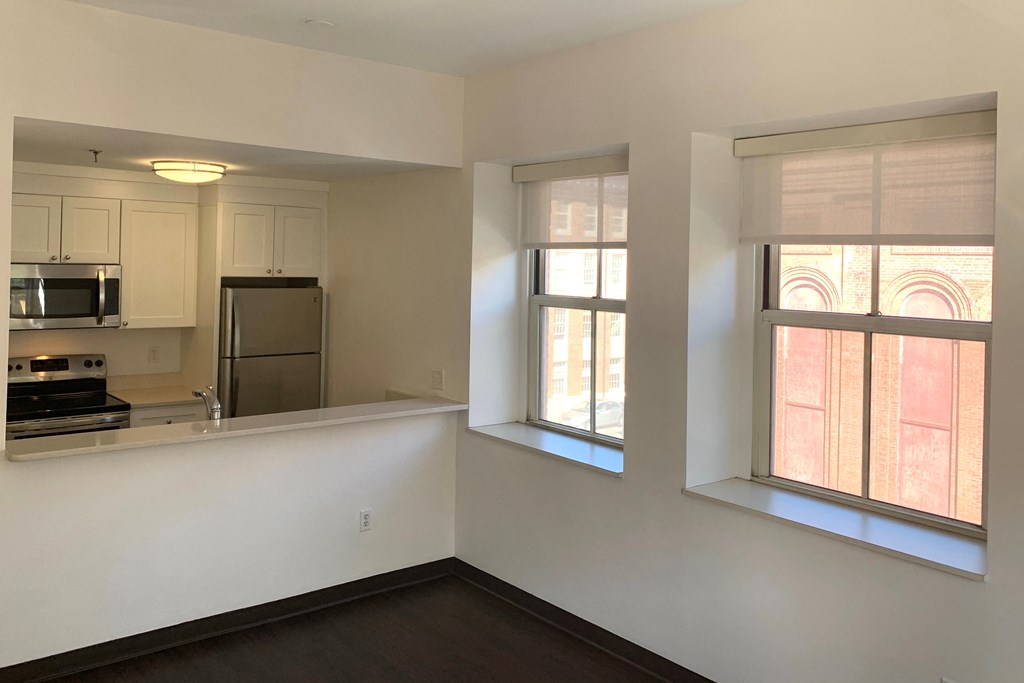 an empty living room with two windows and a kitchen
