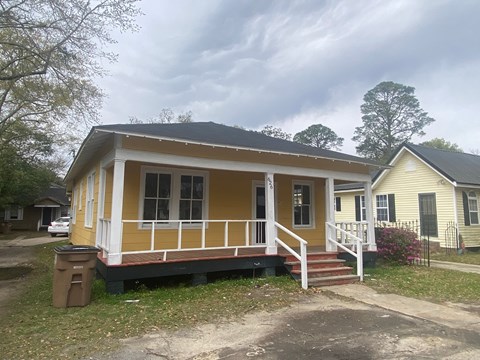 A yellow house with a white porch and a brown trash can in front of it.