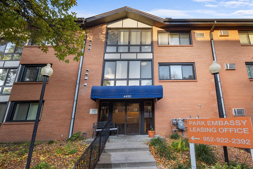 The image shows the front of a brick building with a sign that reads "PARK EMBASSY LEASING OFFICE" and a phone number.