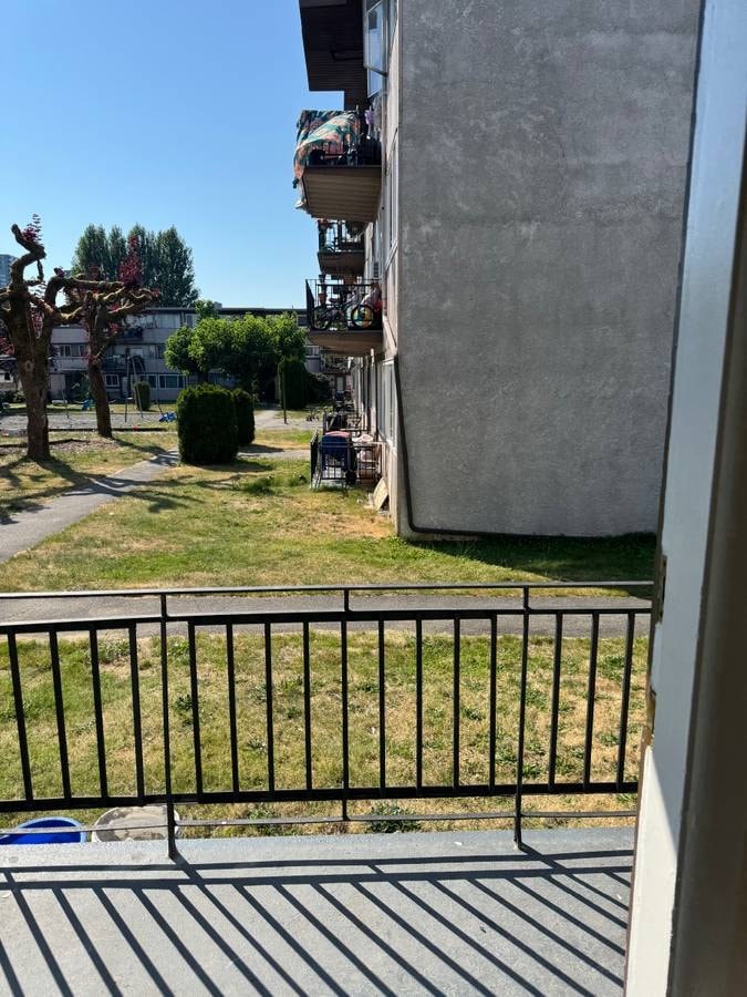 A balcony with a black railing overlooks a grassy area with a tree and a building in the distance.