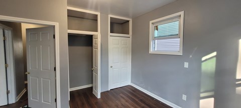 A room with a white door, a window with blinds, and a wooden floor.