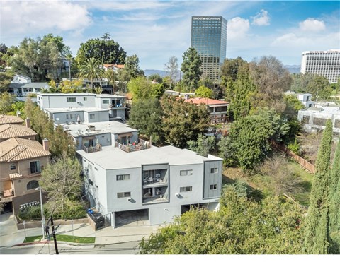 A modern white house with a balcony is surrounded by trees and other houses.