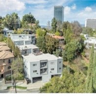 A modern white house is surrounded by trees and other houses.