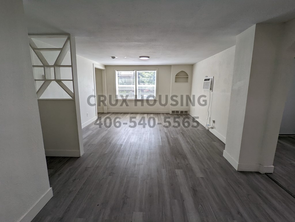 A spacious room with wood flooring and a white wall with a window.