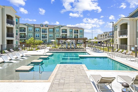 A large swimming pool surrounded by lounge chairs and apartment buildings.