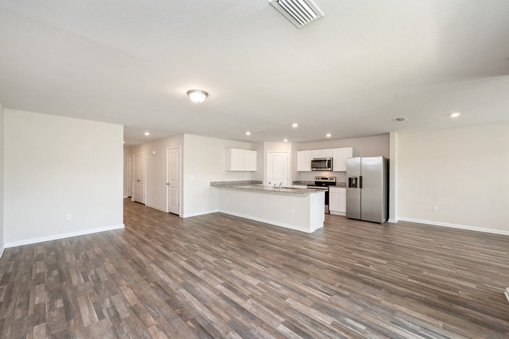 an empty living room with a kitchen with a stainless steel refrigerator