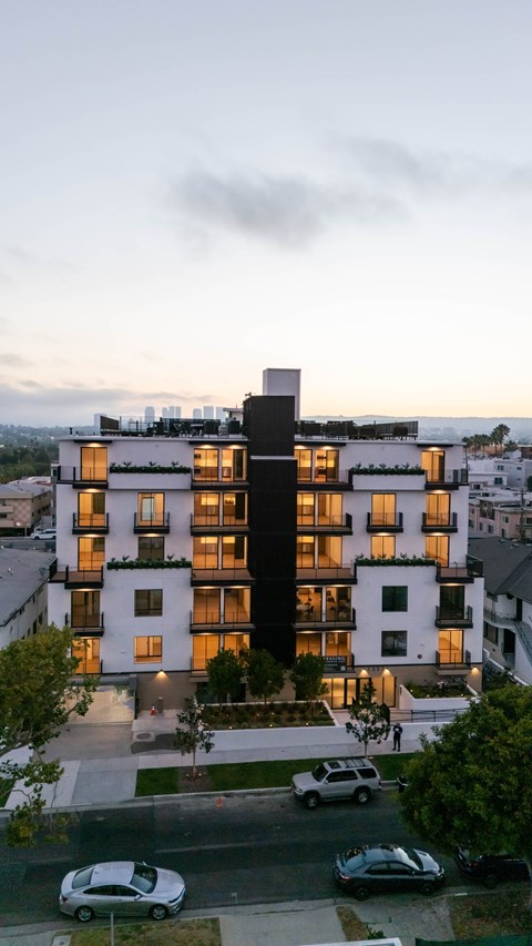 A modern multi-story residential building with balconies and large windows is illuminated from within, with cars parked in front and a cloudy sky above.
