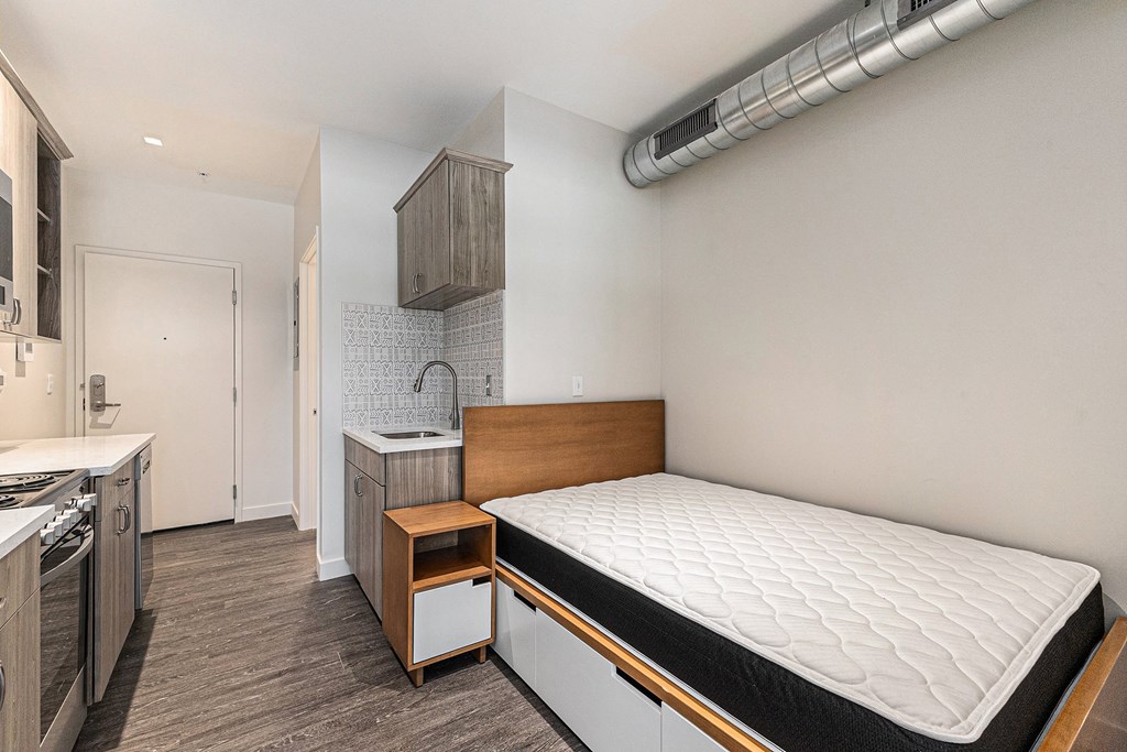 A bedroom with a bed, nightstand, and a kitchen area.