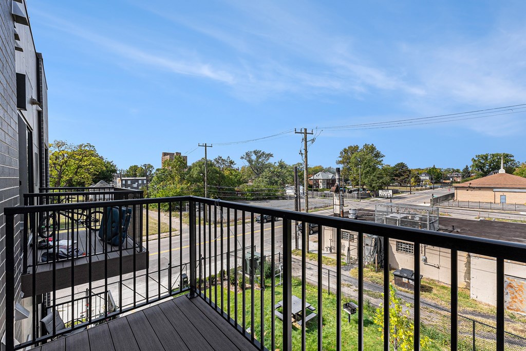 A balcony with a black railing overlooks a parking lot and some buildings.