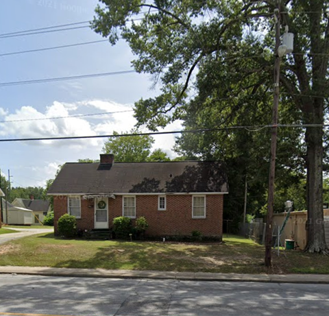 A small brick house with a white door and windows is surrounded by greenery.