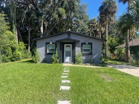 A small house with a grey roof and a white door is surrounded by greenery.