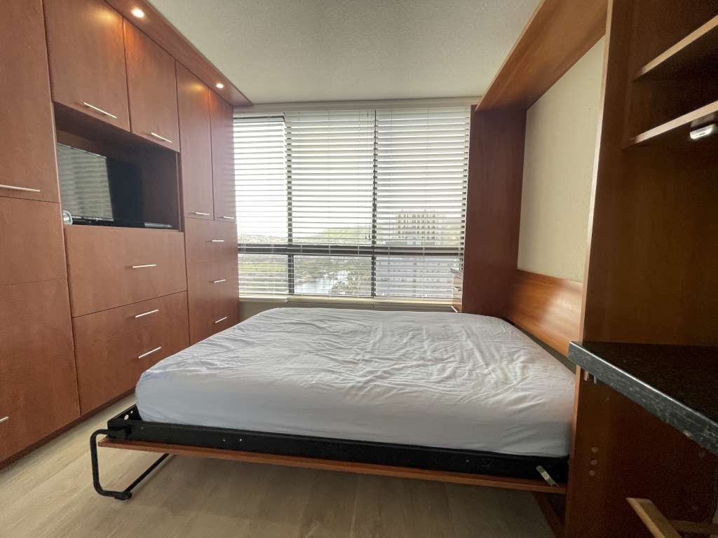 A bed with a white sheet is in a room with wooden cabinets and a window with blinds.