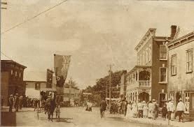 A black and white photo of a street with people and buildings.